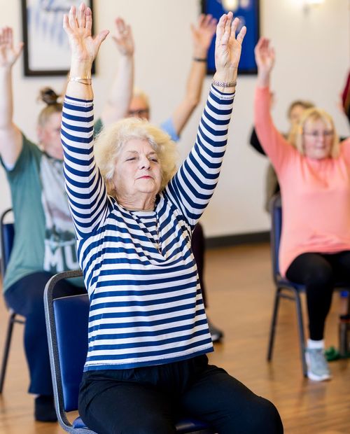 A woman in a seated exercise class reaching both arms overhead.